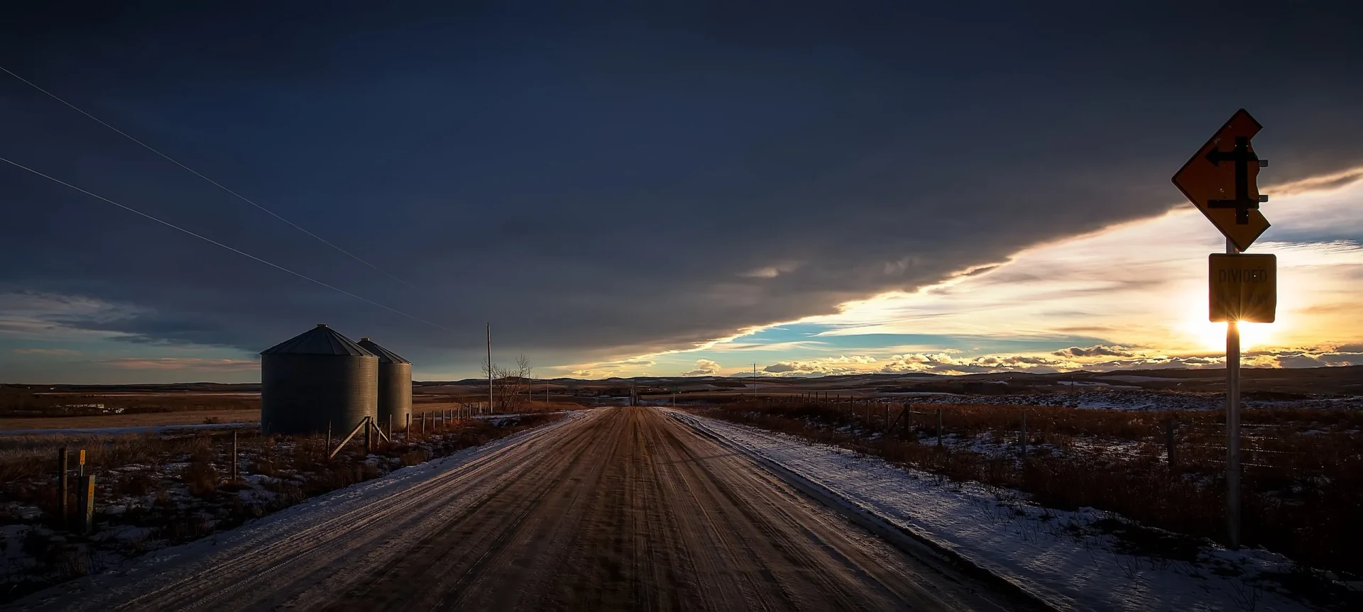 Snow-covered forest road at dusk under a dramatic evening sky.